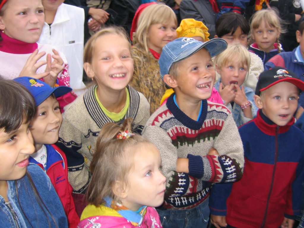 A group of children, dressed in brightly colored clothing, smiling and looking in the same direction. Some are wearing hats and sweaters. The atmosphere seems lively and happy.