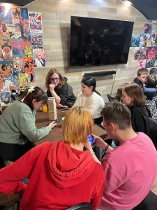 A group of people sit around a table playing Jenga and using their phones in a room decorated with colorful posters and a large wall-mounted TV.