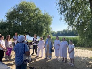 A group of people, some in white robes, stand outdoors near a lake with trees in the background, as others look on.