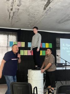 A boy stands on a white drum supported by a man, while another man speaks nearby. Colorful letters are on the wall and a presentation is displayed on a screen.