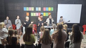 A group of people perform with microphones, keyboard, and guitar onstage in front of an audience of children, with a colorful sign and projection screen behind them.