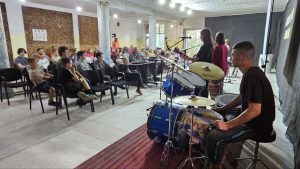 A small audience sits in chairs facing a stage where musicians are performing with drums, microphones, and other instruments in a brightly lit indoor space.