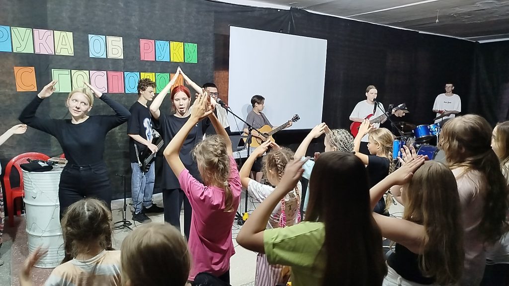 A band performs on stage while a group of children in the audience clap and raise their hands. Colorful letters decorate the wall behind the performers.