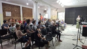 A group of people, including children and adults, sit on chairs facing a woman speaking at the front of a room equipped with microphones and a keyboard.