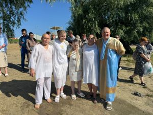 A group of five people in white and blue robes stand together outdoors on a sunny day, with other people and trees visible in the background.