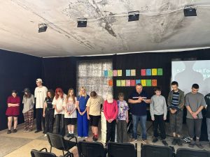 A group of people stands in a line on stage with heads bowed. Behind them, colored paper squares spell words in Cyrillic on a black curtain. Several empty chairs are in the foreground.