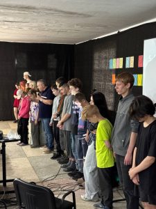 A group of young people and adults stand in a line indoors, heads bowed, in front of a black curtain with colorful letters on the wall.