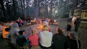 A large group of people, including children and adults, sit in a circle around a campfire in a wooded area near a cabin at dusk.