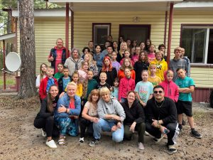 A large group of children and adults poses for a photo outside a yellow building with a porch and satellite dish, surrounded by trees.