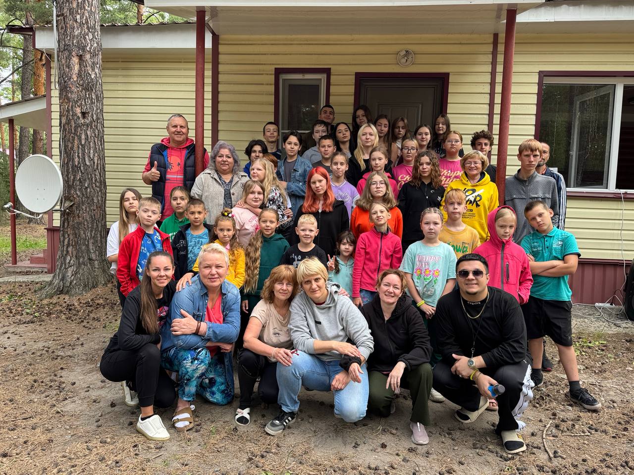 A large group of children and adults poses for a photo outside a yellow house with brown trim, surrounded by trees.