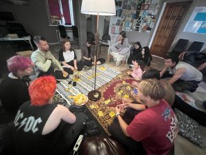 A group of people sit in a circle on the floor around a board game and snacks in a cozy, well-lit room.