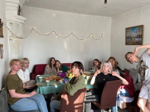 A group of people sit around a dining table with food and drinks, posing for the camera in a well-lit room decorated with string lights.