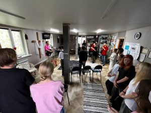 A group of people stands in a room facing a person with a guitar near a microphone, with chairs arranged in rows and various decorations on the walls.