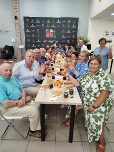 A group of people sitting around a long table with food and drinks, smiling and waving towards the camera in a well-lit indoor setting.