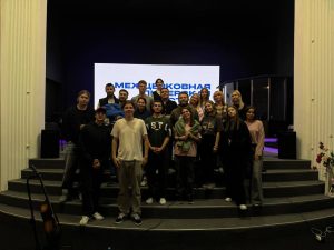 A group of people pose for a photo on indoor steps in front of a large screen displaying text in Russian. Musical instruments and stage equipment are visible.