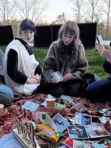 Two women sit on a picnic blanket outdoors, looking at printed photos. Snacks, cups, and various photos are spread out on the blanket, and trees are visible in the background.