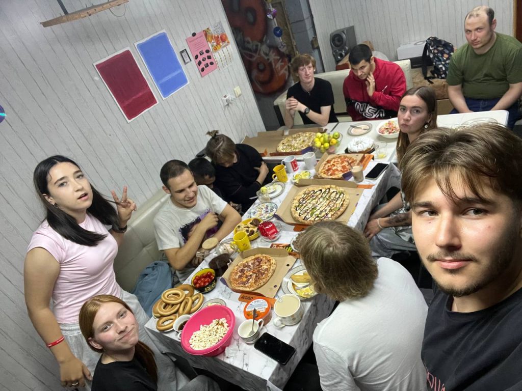 A group of people sit and stand around a table with pizza, snacks, and drinks in a casual indoor setting, posing for a photo together.