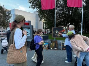 A group of people stands outdoors, observing and discussing framed paintings displayed on the ground near flagpoles and trees.