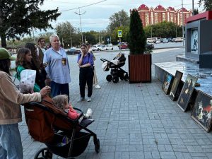 A group of people, including a child in a stroller, look at framed pictures arranged on the ground near a city sidewalk.