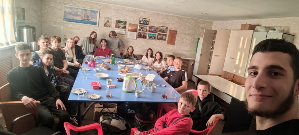 A group of people, mostly children and teenagers, are seated around a blue table with food and drinks in a brightly lit room. One person in the foreground is taking a selfie.