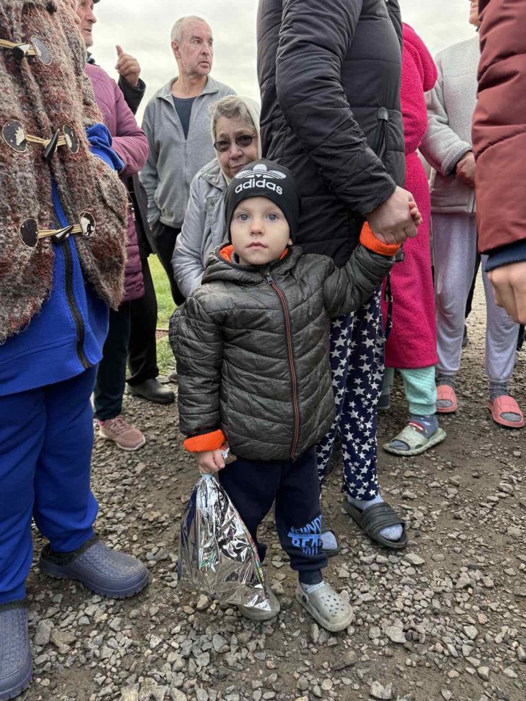 A young child holds an adult’s hand while standing among a group of people outdoors. The child wears a jacket and hat, holding a shiny object. The ground is gravel.