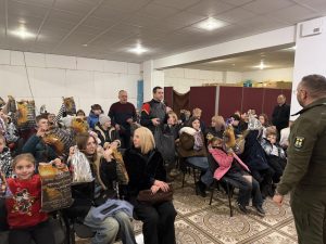 A group of children and adults sit indoors, holding gift bags, while a man in uniform stands and addresses them.