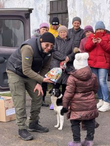 A man hands a gift bag to a child wearing a winter coat and hat, while a dog stands between them. Several people in winter clothing watch and smile in the background.