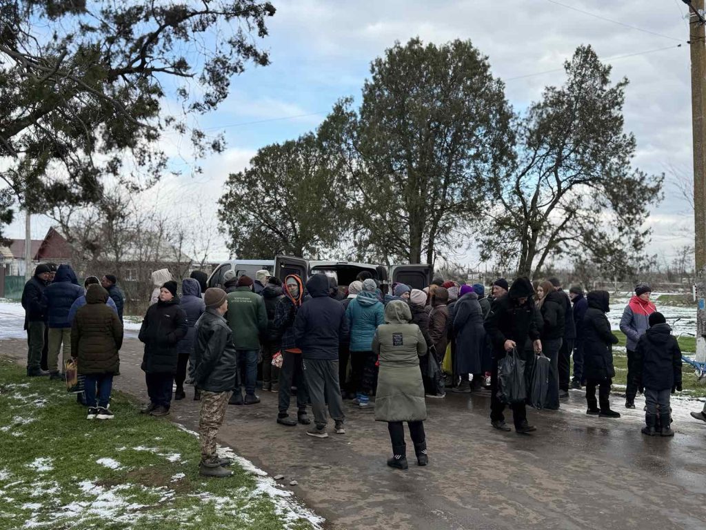 A group of people, many wearing winter clothing, gather around vans on a cold, overcast day with patches of snow on the ground.