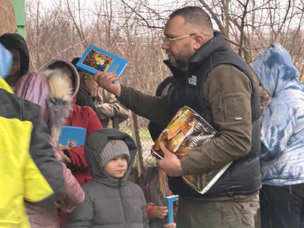 A man distributes books to a group of children dressed in winter clothing outdoors on a cloudy day.