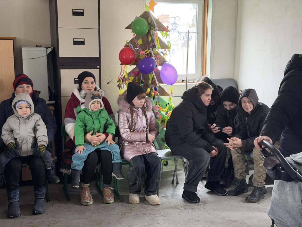 A group of children and adults in winter clothing sit on benches in a room with a small decorated tree and balloons by the window.