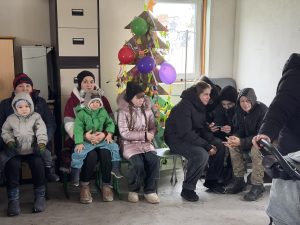 A group of children and adults in winter clothing sit on benches in a room with a small decorated tree and balloons by the window.