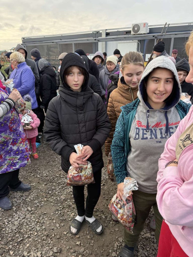 A group of people, including children and adults in warm clothing, stand outdoors on gravel ground holding bags with various designs.