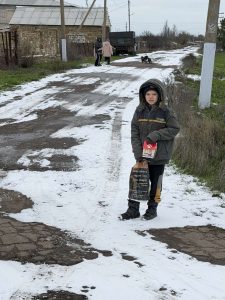 A boy in winter clothing stands on a partially snow-covered rural road holding a bag and a snack, while three people and a dog are visible in the distance.