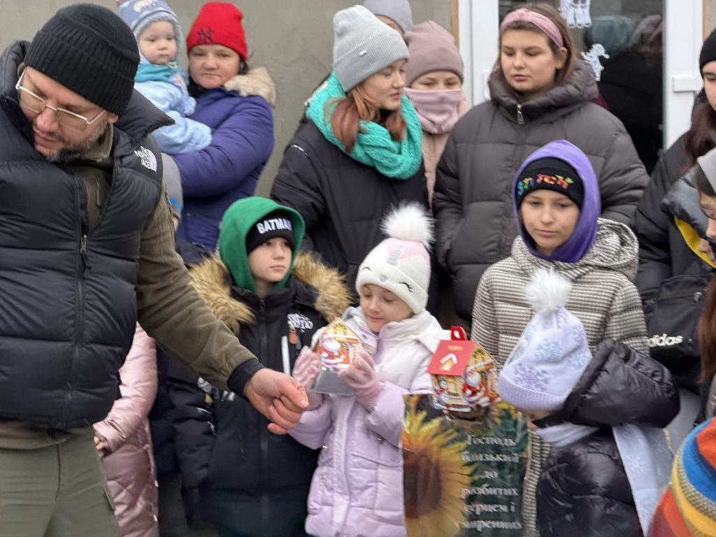 A group of adults and children dressed in winter clothing stand outside. Two children in front hold packaged gifts and look at them.