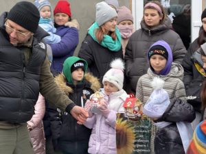 A group of adults and children dressed in winter clothing stand outside. Two children in front hold packaged gifts and look at them.