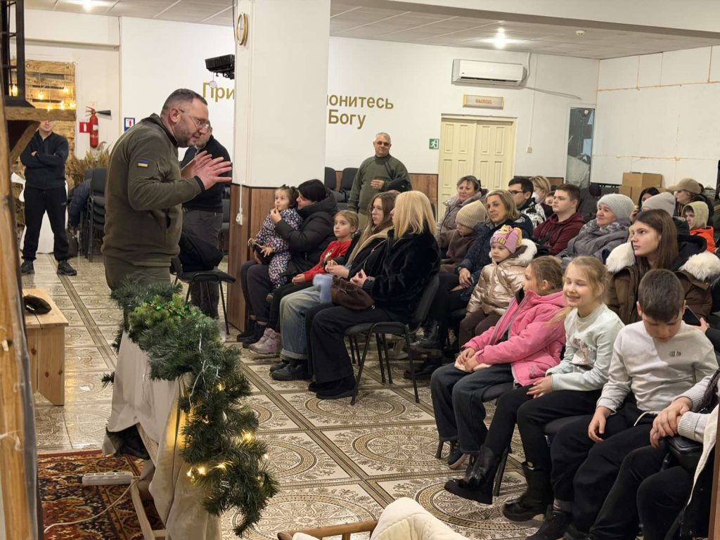 A man speaks to a seated audience of adults and children in a decorated indoor setting with holiday greenery and religious wall text in the background.