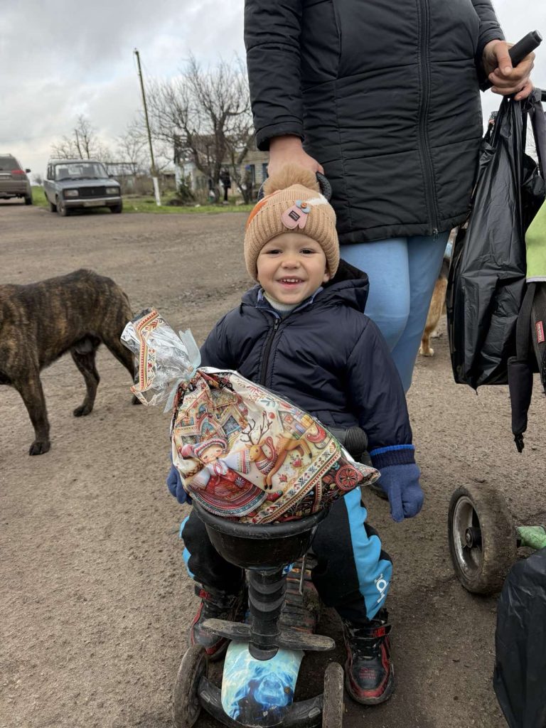 A young child sits on a scooter holding a decorated package, smiling, while an adult stands behind, holding bags. A dog and vehicles are visible in the background.