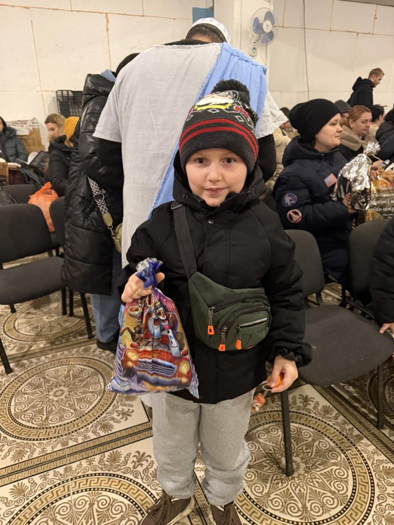 A child in a winter coat and hat stands indoors, holding a gift bag, with other people seated in the background.