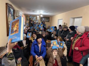 A group of children and adults in a room hold up colorful books, smiling and posing for the camera; some are seated while others stand in winter clothing.
