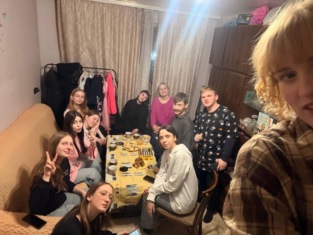 A group of twelve people sit and stand around a table set with food and drinks in a living room, posing for a photo.
