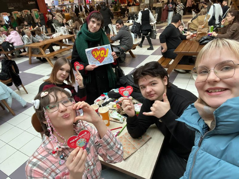 Five young people at a crowded food court sit around a table holding decorated heart-shaped items, smiling and posing for a selfie.