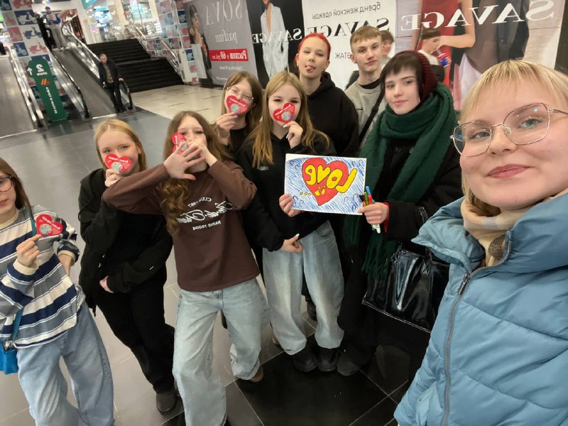 A group of young people pose in a mall, some holding heart-shaped items and one holding a "Love" sign. Escalators and storefronts are visible in the background.