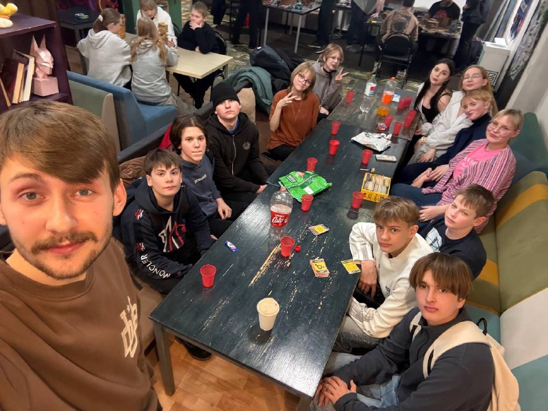 A group of teenagers and adults sit around a table with snacks, drinks, and playing cards in a casual indoor setting.