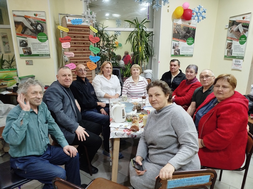 A group of ten adults sit around a table with food and drinks in a decorated indoor setting, looking at the camera.