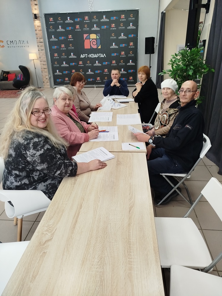 Eight adults sit around a rectangular table with papers and pens, appearing to participate in a meeting at an indoor venue with a branded backdrop.