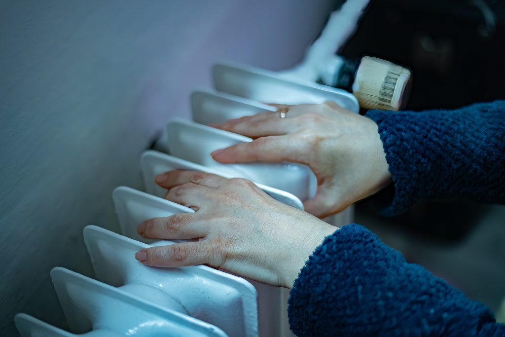 Two hands in a blue fleece sweater are held above a white radiator, suggesting an attempt to warm up.
